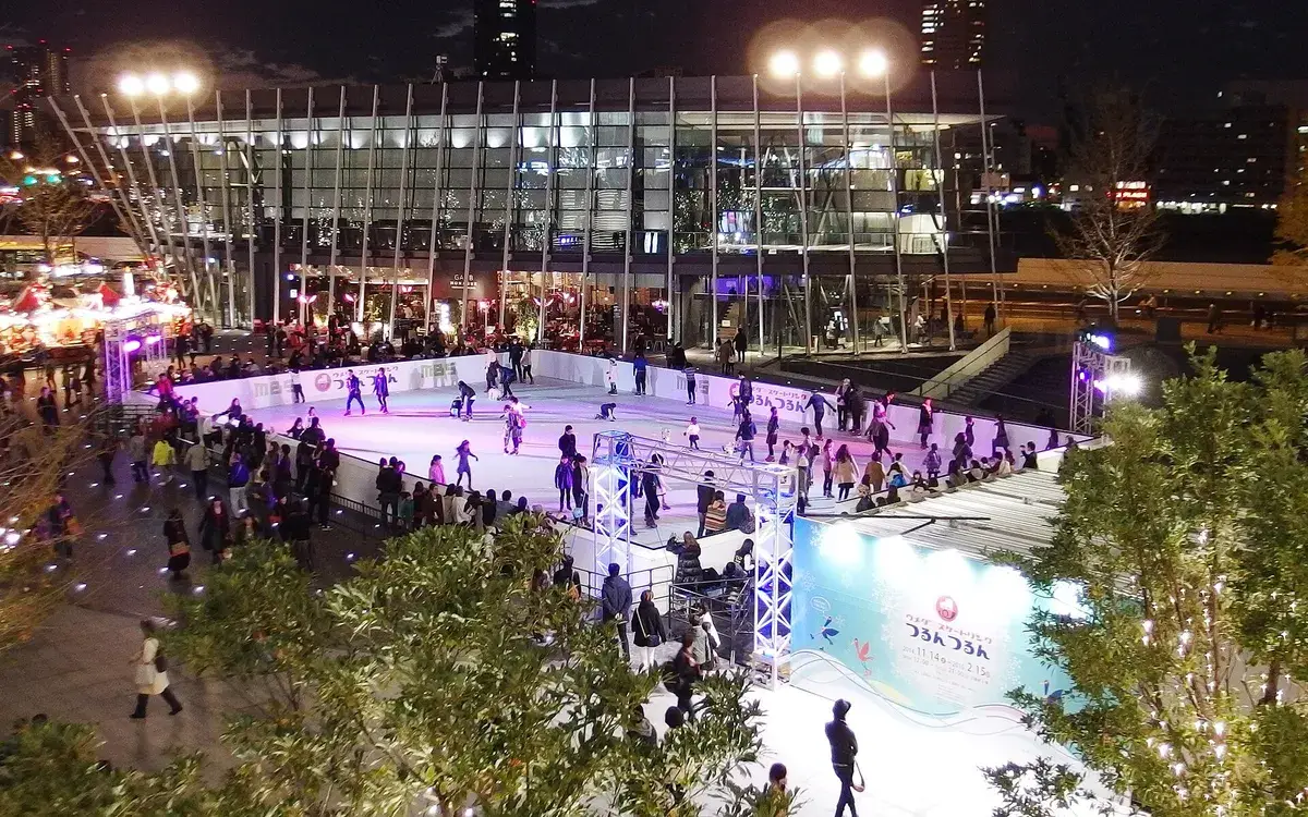 An outdoor skating rink at night in a city, skaters beneath tall buildings and lights