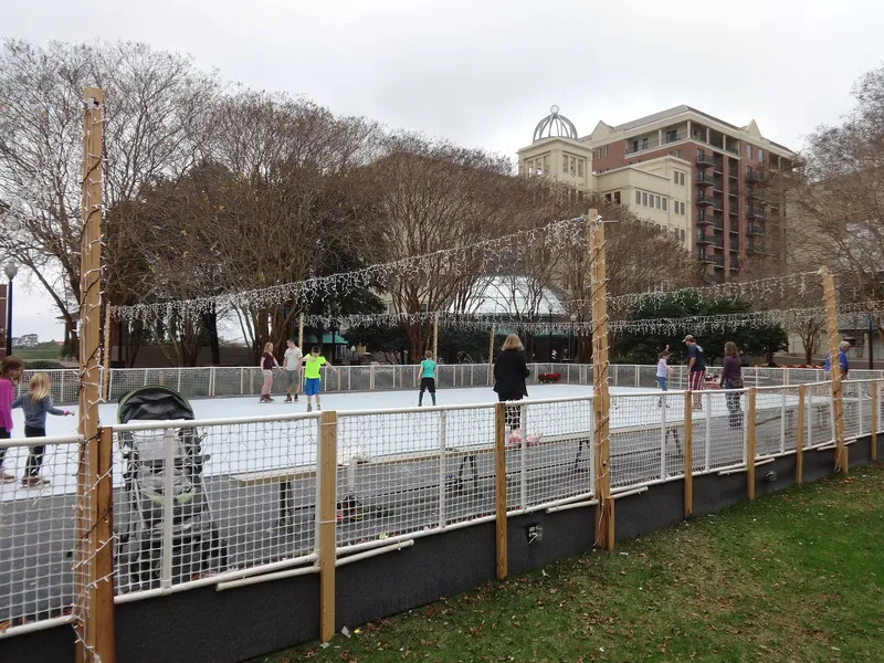 Children skating on an outdoor rink strung with Christmas lights, a stroller beside the rink fence in daytime