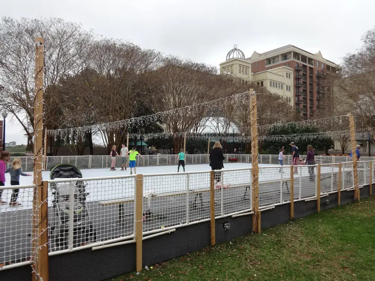 Children skating on an outdoor rink strung with Christmas lights, a stroller beside the rink fence in daytime