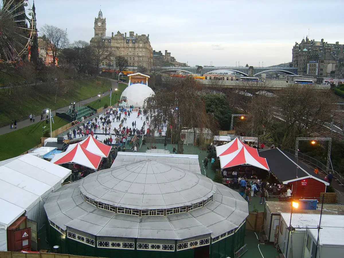 Skaters on an outdoor rink beside a Christmas market, stalls and lights nearby