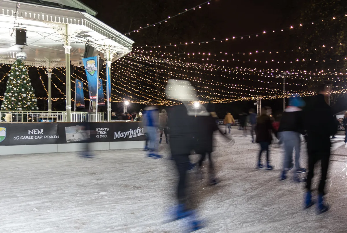 Skaters on an outdoor rink at a night winter festival, bandstand, Christmas tree and strings of lights around the ice