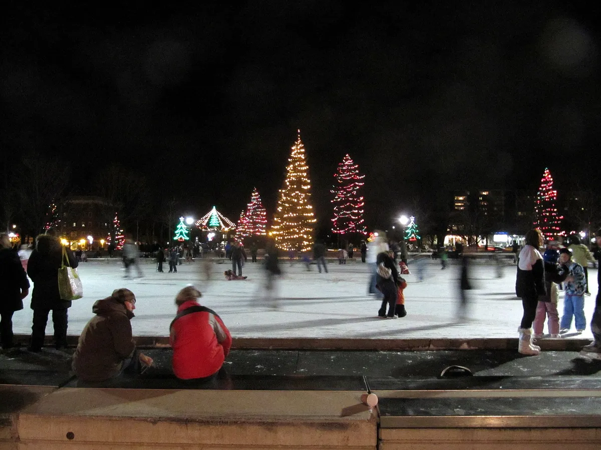 Skaters on an outdoor rink in a city park, trees and snow around
