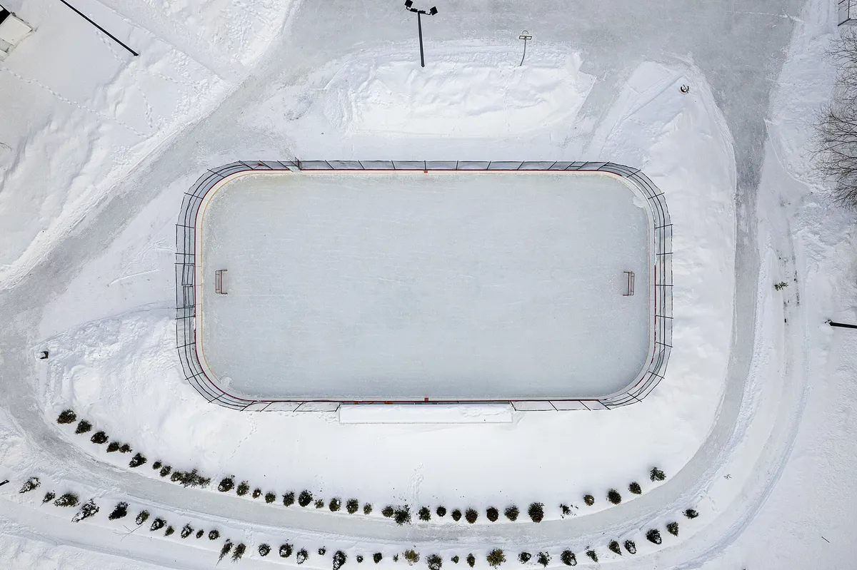 An outdoor ice rink seen from above, ringed by snow and trees