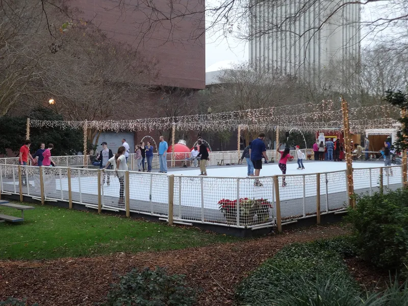 Skaters on a large outdoor rink strung with Christmas lights, a giant red ornament beside the rink in daytime