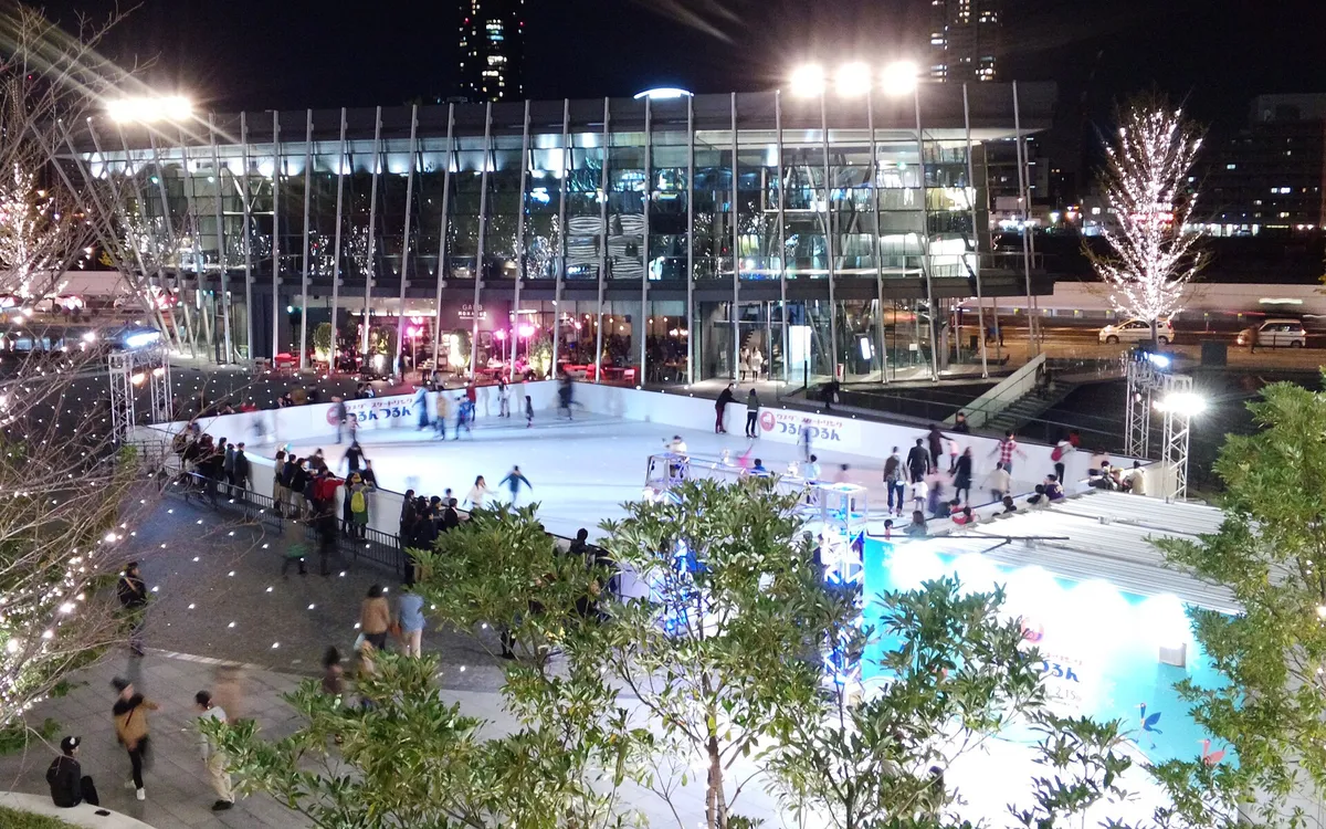 Skaters on an outdoor city rink at night, lit buildings and a tall tower behind