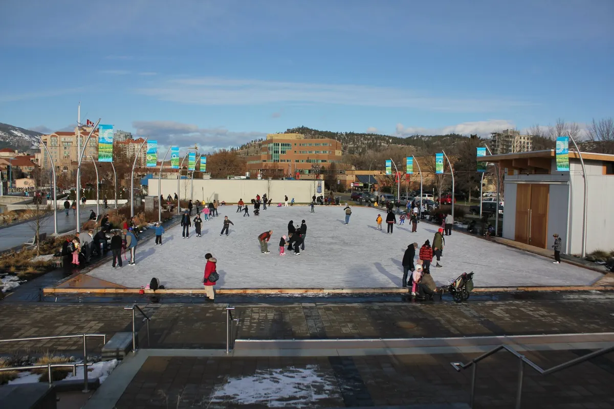 Families skating on a sunny outdoor park rink, mountains and city buildings behind